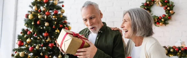 Щаслива зріла жінка дивиться на здивованого чоловіка з різдвяним подарунком біля прикрашеної ялинки, банер — стокове фото