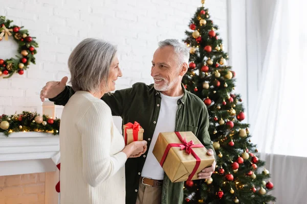 Веселая зрелая женщина обменивается рождественскими подарками со счастливым мужем возле украшенной ели — стоковое фото