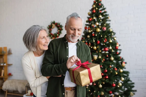 Улыбающаяся женщина обнимает взрослого бородатого мужа с подарком на Рождество — стоковое фото
