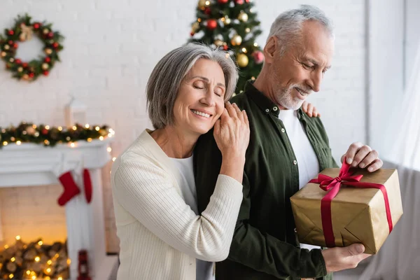 Усміхнена жінка з закритими очима обіймає зрілого бородатого чоловіка з різдвяним подарунком — стокове фото