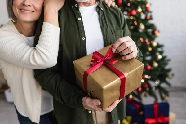Cropped view of happy middle aged woman hugging husband with christmas present — Stock Photo