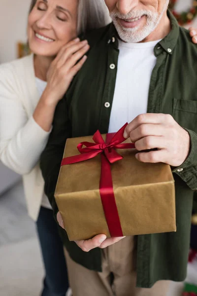 Vista cortada de alegre mulher de meia idade abraçando marido barbudo com presente de Natal — Fotografia de Stock