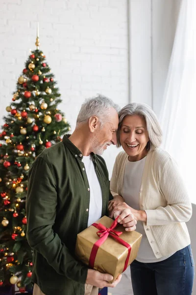 Бородатый мужчина средних лет держит подарок и обнимает счастливую жену возле рождественской елки — стоковое фото