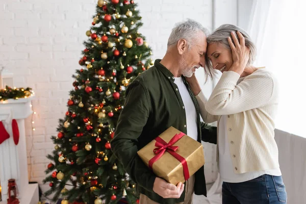 Бородатий чоловік середнього віку тримає подарунок і обіймається з усміхненою дружиною біля ялинки — стокове фото