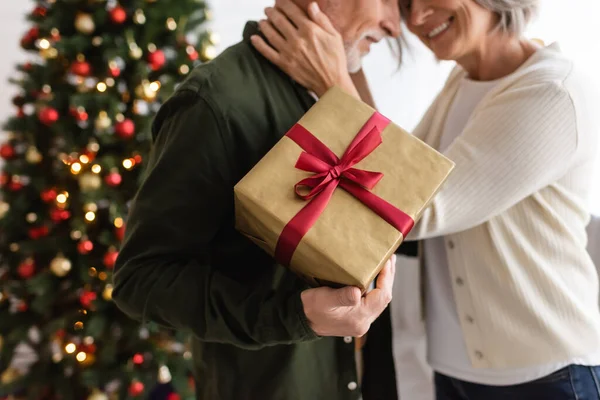 Vista recortada del hombre barbudo de mediana edad sosteniendo presente y abrazándose con esposa feliz cerca del árbol de Navidad - foto de stock