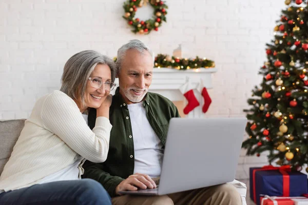 Щаслива пара середнього віку дивиться фільм на ноутбуці біля прикрашеної ялинки — стокове фото