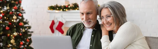 Счастливая пара средних лет смотреть фильм на ноутбуке рядом с украшенной елкой, баннер — стоковое фото