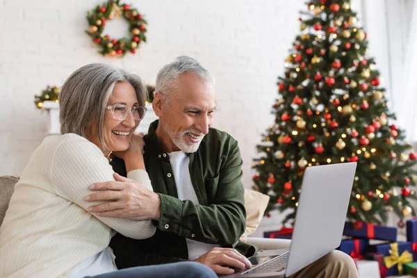 Усміхнена пара середнього віку дивиться комедійний фільм на ноутбуці біля прикрашеної ялинки — стокове фото