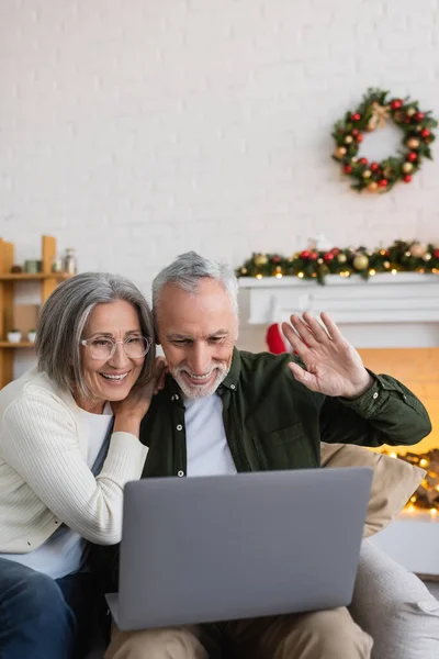 Улыбающийся мужчина средних лет машет рукой возле жены во время видеозвонка на ноутбуке во время Рождества — стоковое фото