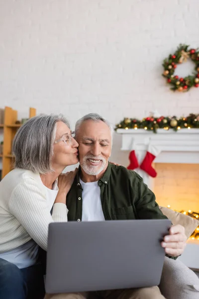 Smiling middle aged woman kissing cheek of husband during having video call on laptop on christmas day — Stock Photo