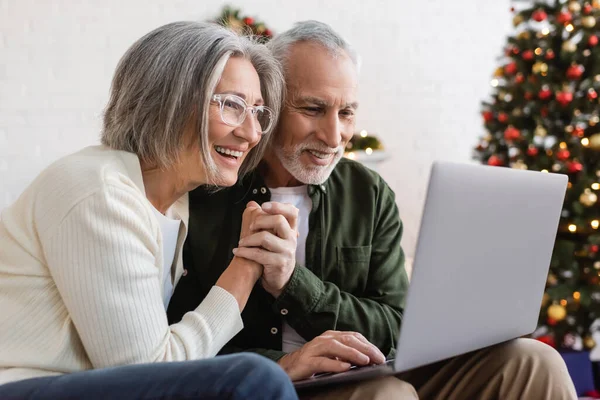 Улыбающаяся пара средних лет, держащаяся за руки и имеющая видеозвонок на ноутбуке во время Рождества — стоковое фото