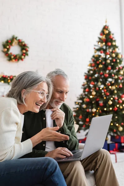 Smiling middle aged woman gesturing near husband and having video call on laptop during christmas — Stock Photo