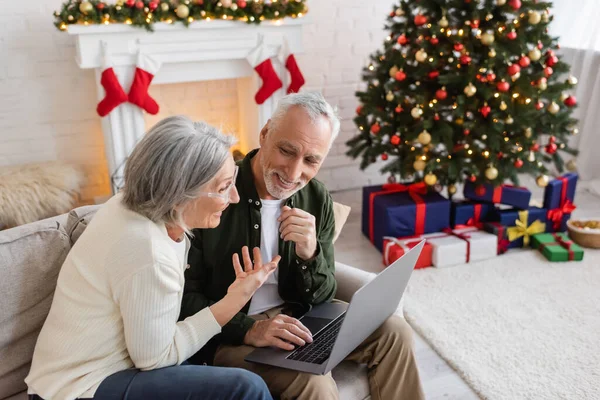 Smiling middle aged woman pointing at happy husband and having video call on laptop during christmas — Stock Photo