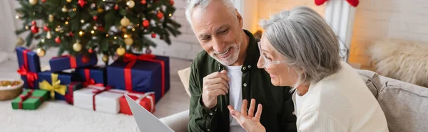 Усміхнена жінка середнього віку вказує на щасливого чоловіка і має відеодзвінок на ноутбук під час Різдва, банер — стокове фото