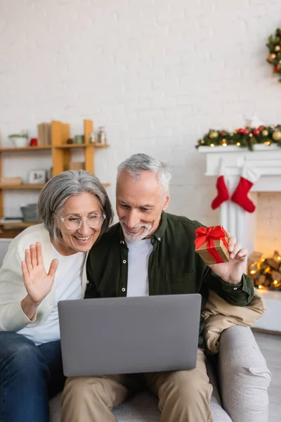 Sorrindo mulher de meia idade acenando mão perto do marido e tendo chamada de vídeo no laptop durante o Natal — Fotografia de Stock