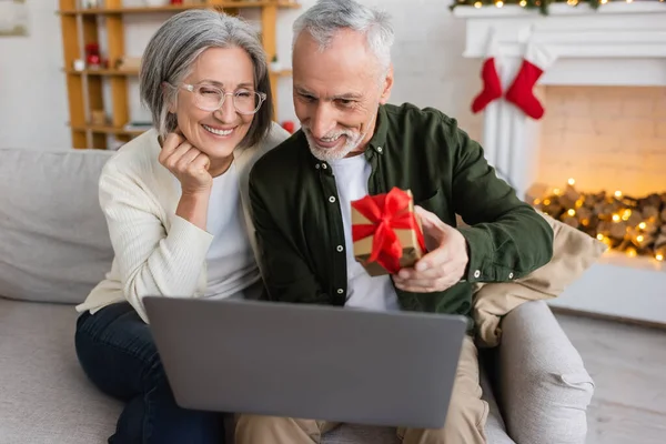 Glücklicher Mann mittleren Alters mit Weihnachtsgeschenk in der Nähe von Frau und Laptop während eines Videogesprächs — Stockfoto