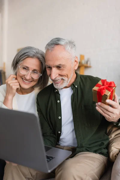 Fröhlicher Mann mittleren Alters hält während Videoanruf Weihnachtsgeschenk in der Nähe seiner Frau — Stockfoto