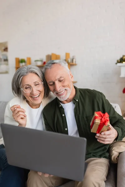 Alegre homem de meia idade segurando Natal presente perto sorrindo esposa durante a chamada de vídeo — Fotografia de Stock