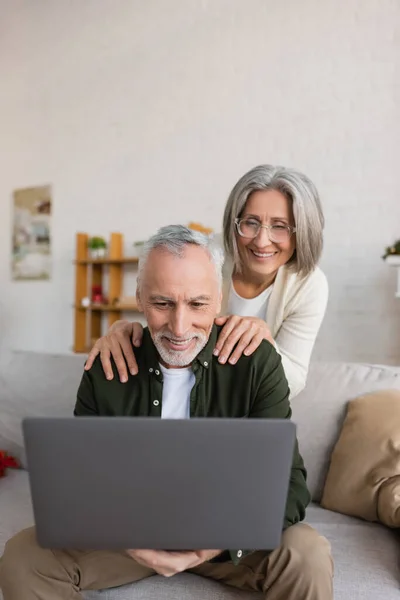Cheerful middle aged woman in glasses hugging husband during video call on laptop — Stock Photo