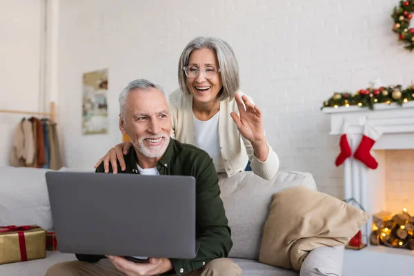 Alegre mulher de meia-idade em óculos acenando mão perto do marido durante chamada de vídeo no dia de Natal — Fotografia de Stock