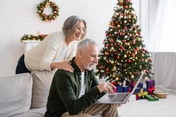Cheerful middle aged woman in glasses hugging husband during video call near christmas tree — Stock Photo