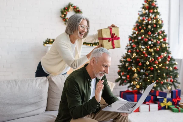 Cheerful middle aged woman in glasses showing christmas present near husband during video call — Stock Photo
