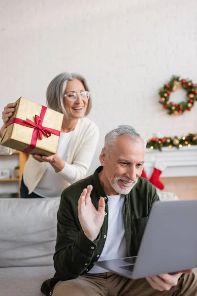 Cheerful middle aged woman in glasses showing christmas present near husband waving hand during video call — Stock Photo