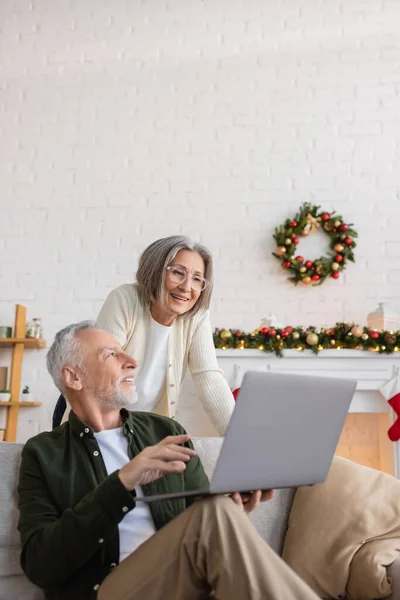Alegre hombre de mediana edad mostrando portátil a la esposa en gafas en el día de Navidad - foto de stock