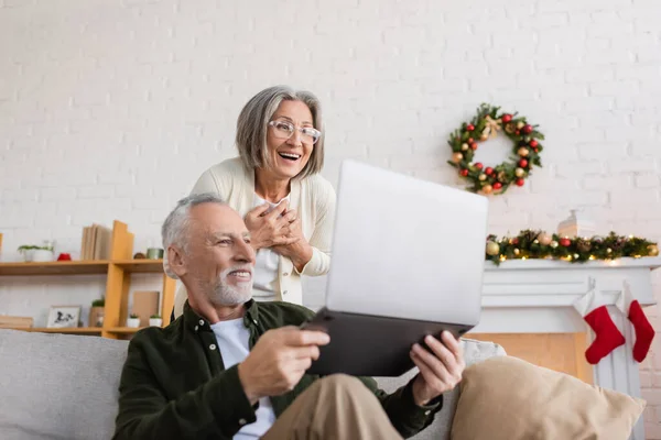 Веселий чоловік середнього віку, що показує ноутбук ледачій дружині в окулярах на Різдво — стокове фото