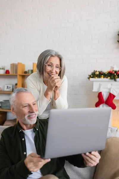 Щасливий чоловік середнього віку, що показує ноутбук лежачій дружині в окулярах на Різдво — стокове фото