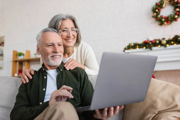 Mujer de mediana edad feliz en gafas abrazando marido y mirando a la computadora portátil durante la videollamada en el día de Navidad - foto de stock