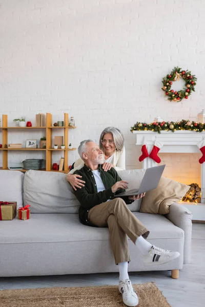 Счастливая женщина средних лет в очках обнимает мужа, сидящего на диване с ноутбуком на Рождество — стоковое фото