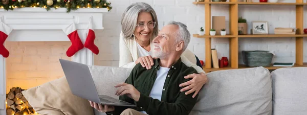 Mujer de mediana edad feliz en gafas abrazando marido con el ordenador portátil en el día de Navidad, bandera - foto de stock