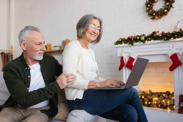 Весела жінка середнього віку в окулярах використовує ноутбук біля щасливого чоловіка під час різдвяних свят — стокове фото