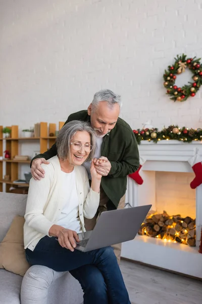Mujer alegre de mediana edad en gafas usando el ordenador portátil y tomados de la mano con el marido feliz durante las vacaciones de Navidad - foto de stock