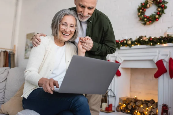 Счастливая женщина средних лет в очках с помощью ноутбука и держаться за руки с мужем во время рождественских праздников — стоковое фото