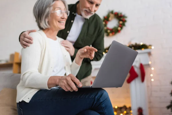 Glückliche Frau mittleren Alters in Brille zeigt auf Laptop in der Nähe glücklicher Ehemann während der Weihnachtsfeiertage — Stockfoto