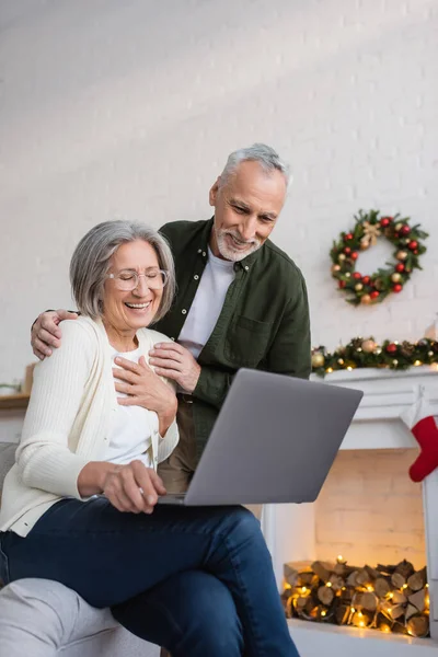 Feliz de mediana edad marido y esposa riendo cerca de la computadora portátil durante las vacaciones de Navidad - foto de stock