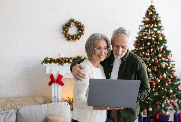 Marido y esposa de mediana edad feliz mirando el portátil cerca del árbol de Navidad decorado - foto de stock