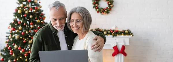 Glückliches Ehepaar mittleren Alters blickt auf Laptop neben geschmücktem Weihnachtsbaum, Banner — Stockfoto