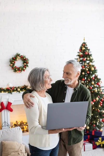 Happy middle aged husband hugging wife holding laptop near decorated christmas tree — Stock Photo