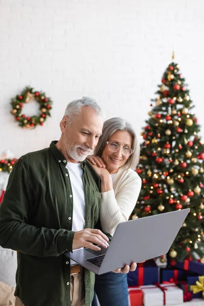 Весёлые муж и жена средних лет смотрят на ноутбук рядом с украшенной елкой — стоковое фото