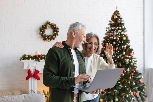 Alegre homem de meia-idade olhando esposa acenando mão durante vídeo chat perto de árvore de natal decorada — Fotografia de Stock