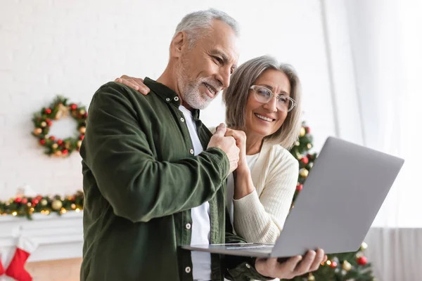 Весёлые муж и жена средних лет держатся за руки и смотрят на ноутбук — стоковое фото