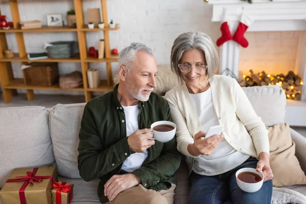 Vue grand angle de la femme mûre dans des lunettes en utilisant un smartphone et tenant tasse près du mari pendant les vacances de Noël — Photo de stock