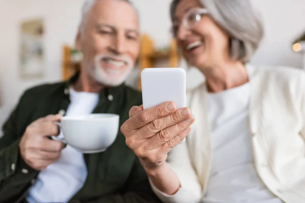 Mujer alegre y madura sosteniendo teléfono inteligente cerca marido feliz con taza de té - foto de stock