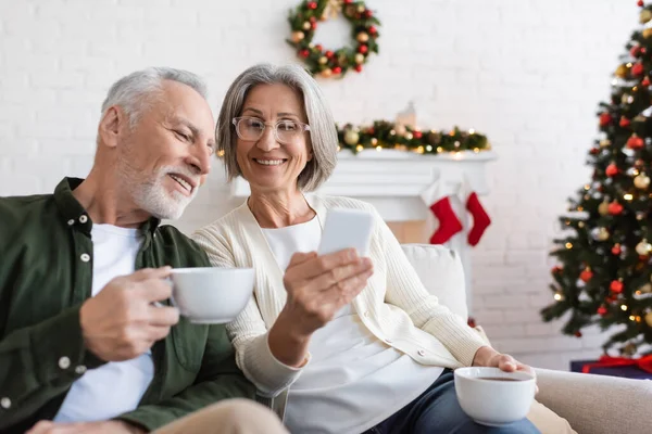 Усміхнена і зріла жінка в окулярах тримає смартфон біля чоловіка з чашкою чаю під час різдвяних канікул — стокове фото