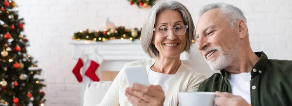 Mujer sonriente y madura en gafas con teléfono inteligente cerca del marido con taza de té, pancarta - foto de stock