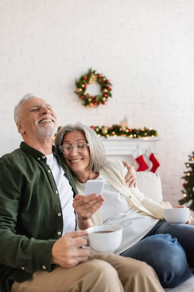 Весела і середнього віку жінка в окулярах тримає смартфон біля щасливого чоловіка з чашкою чаю — стокове фото