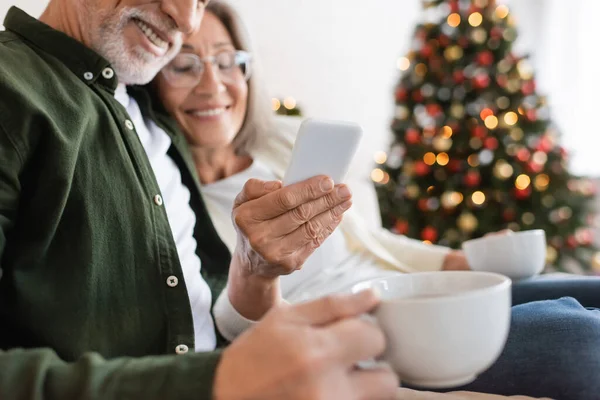 Веселая женщина средних лет в очках держит смартфон рядом с улыбающимся мужем с чашкой чая — стоковое фото